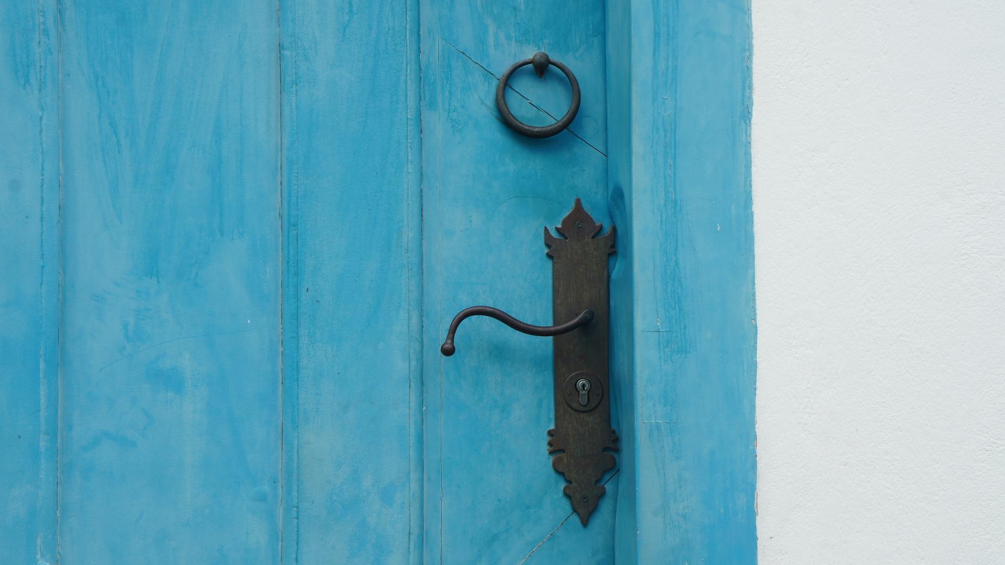 A close-up of a rustic wooden door painted blue, featuring a vintage iron handle and door knocker.